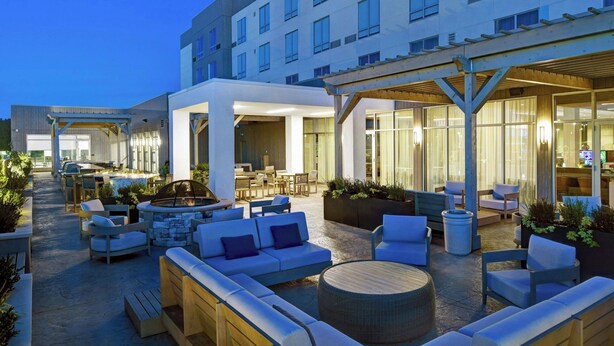 A modern hotel with outdoor seating areas, a pool, and a covered patio. - Courtyard by Marriott Hamilton (Robbinsville)