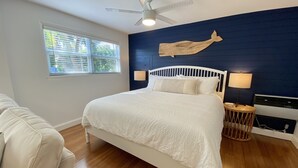 A bedroom with a white bed, a ceiling fan, a window with blinds, a blue accent wall, and a small bedside table with a lamp. - Studio steps from Intracoastal & downtown WPB with bikes (West Palm Beach)
