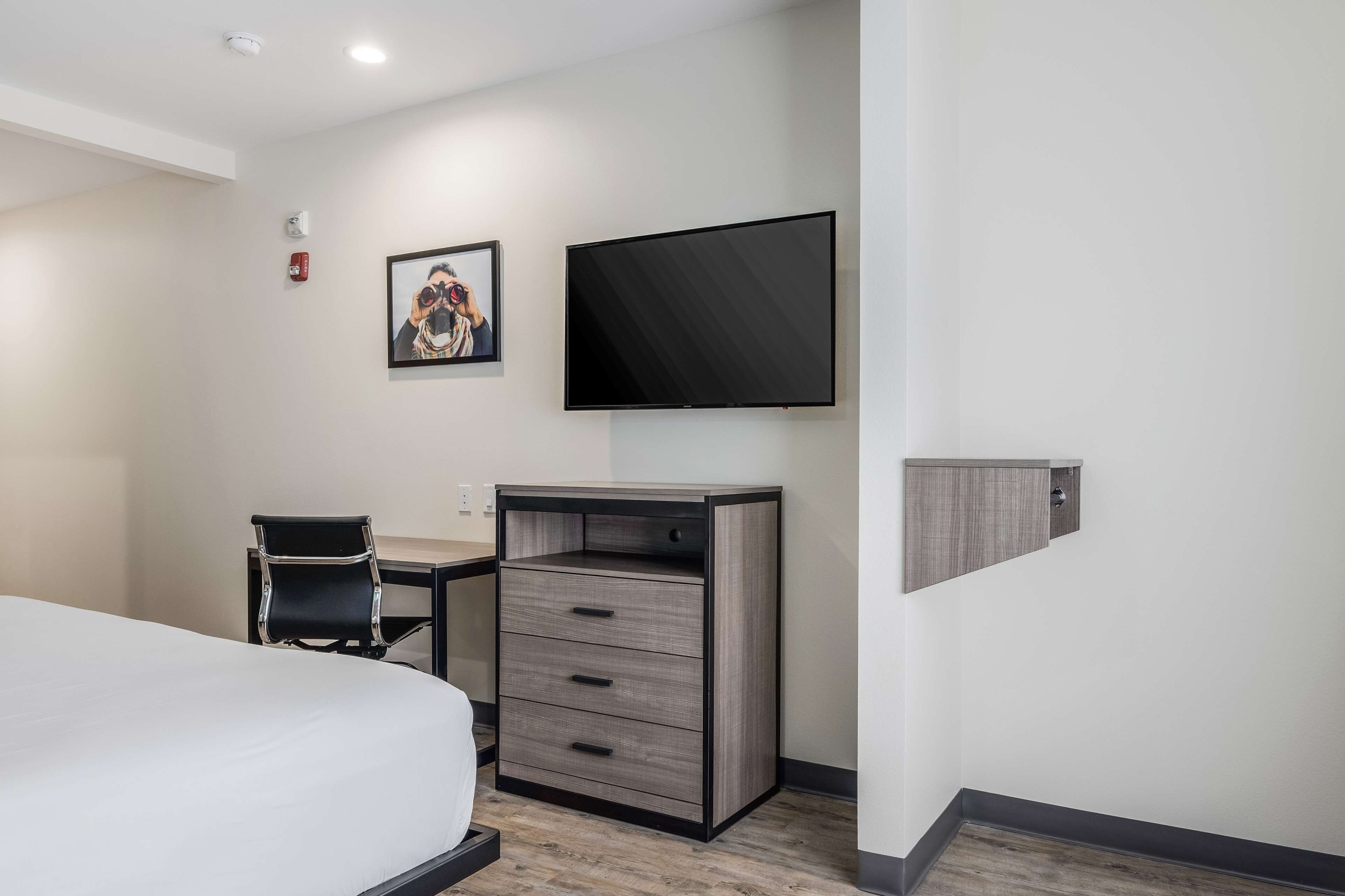 A hotel room with a bed, a desk with drawers, a chair, a flat-screen TV mounted on the wall, and a framed picture.
