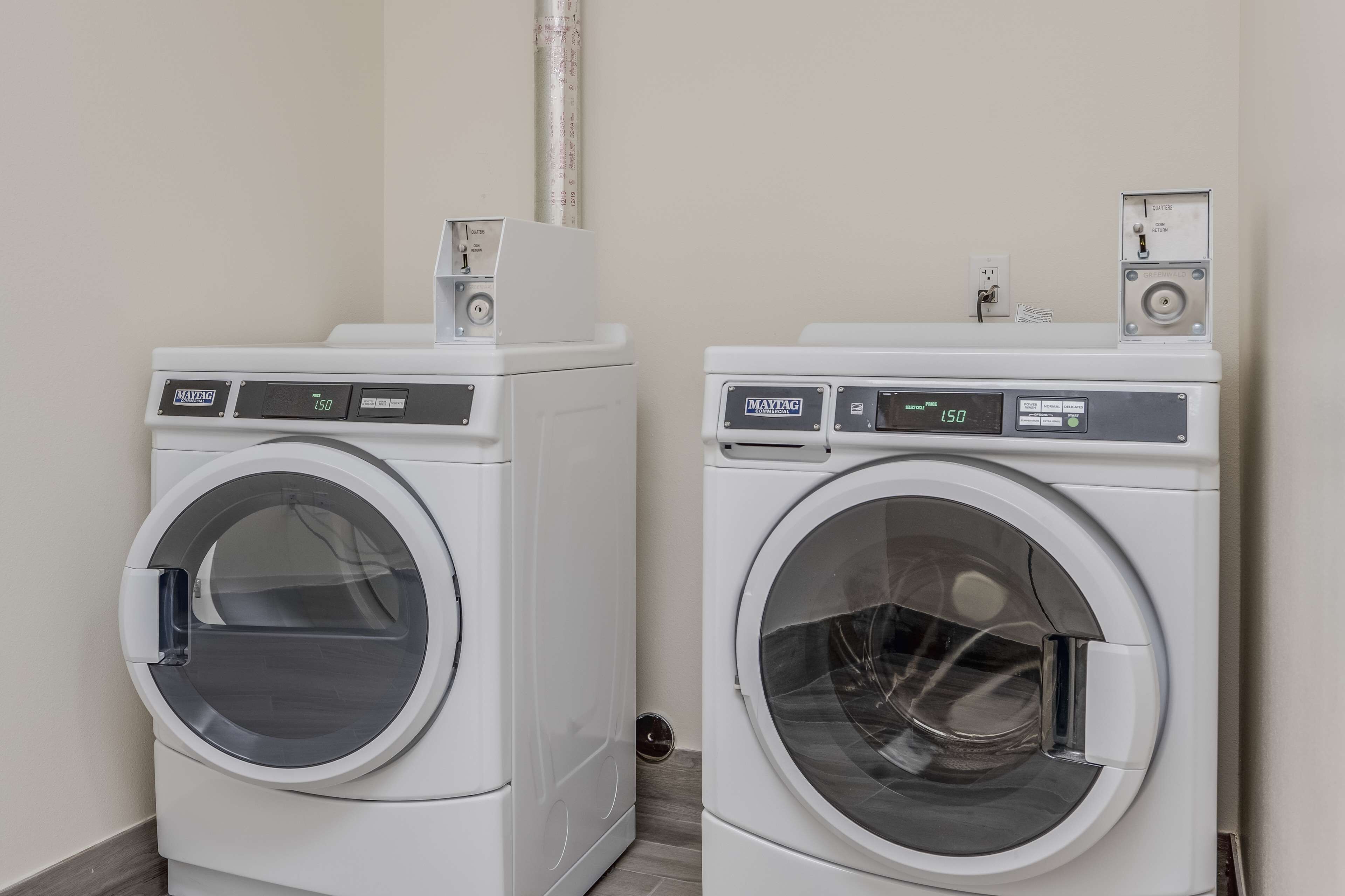 Two white front-loading washing machines in a laundry room with a white wall and a pipe above them.