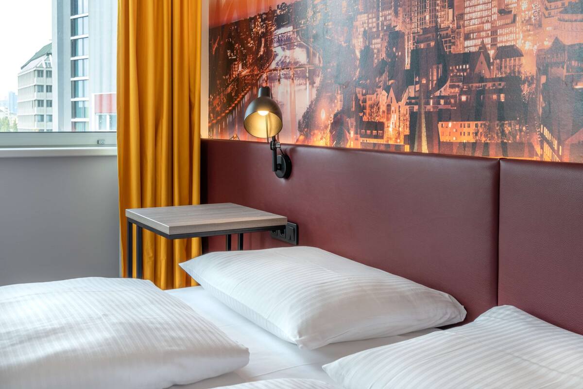 A hotel room with a bed, a nightstand, a lamp, a cityscape mural, and a window with curtains.