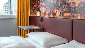 Habitación de hotel con cama, mesita de noche, lámpara, un mural de paisaje urbano y una ventana con cortinas.