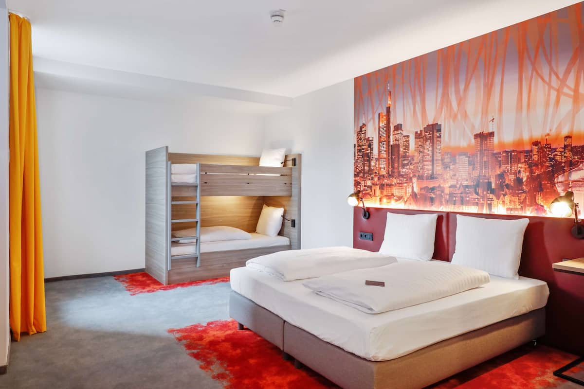 A hotel room with a bed, a bunk bed, a cityscape mural, and a red carpet.