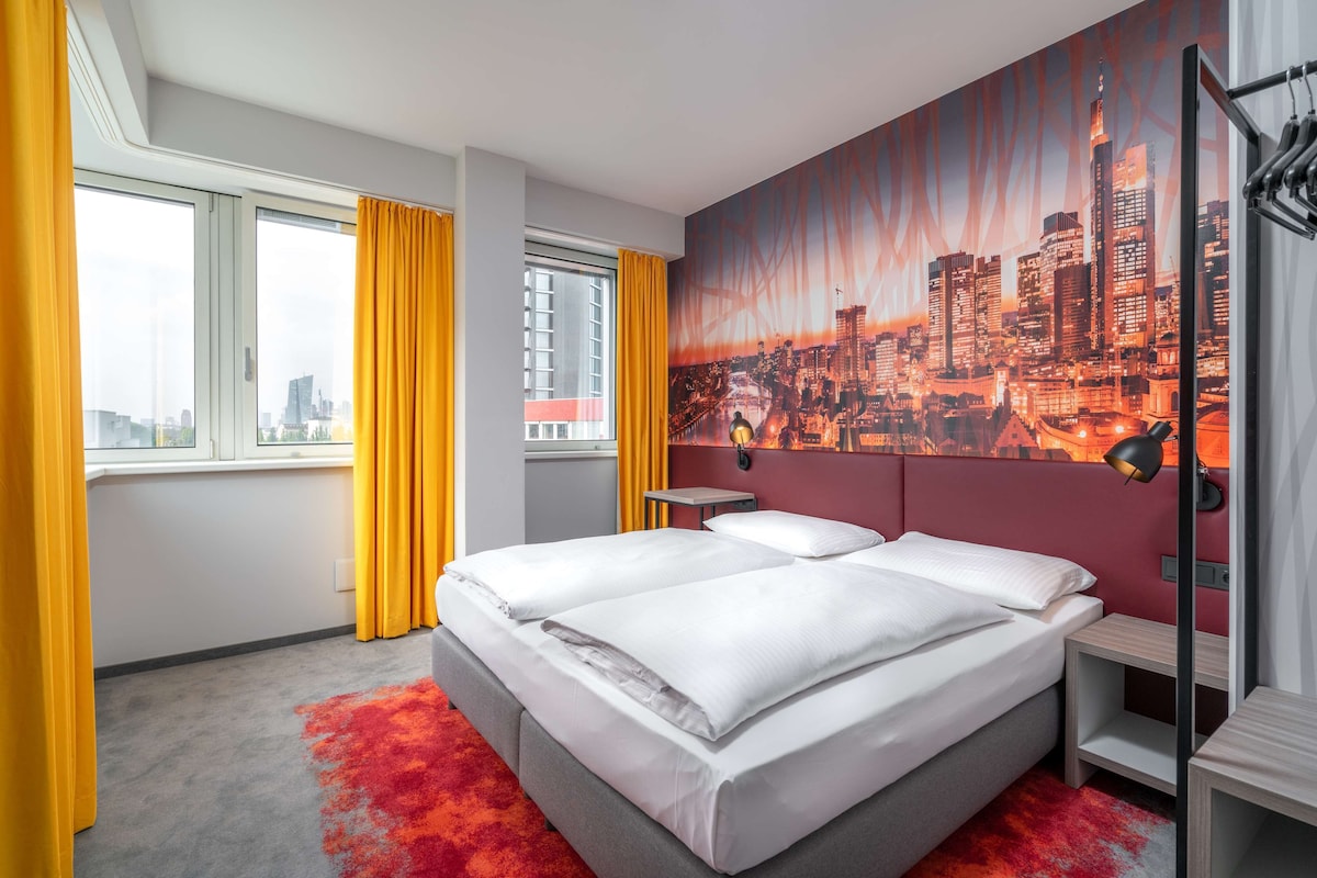 A hotel room with a large bed, a cityscape mural, a window with curtains, and a red carpet.