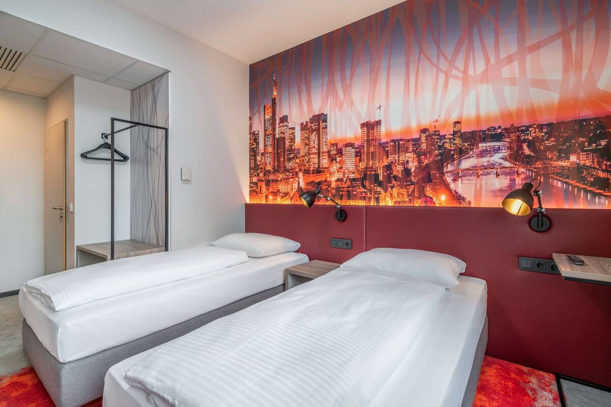 A hotel room with two beds, a cityscape mural, a red accent wall, and a hanging coat rack.