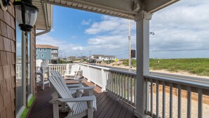 A wooden deck with white rocking chairs, a white railing, and a view of the beach and buildings in the background. - Pet Friendly, Ocean Views, Pool & Hot Tub! (Kitty Hawk)