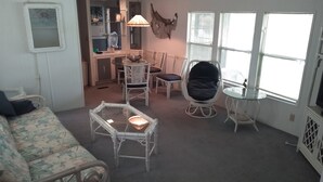 Un salon comprenant un canapé, une table basse avec un plateau en verre, des fauteuils en osier, une table à manger avec des chaises, un coin cuisine et une grande fenêtre avec des stores.