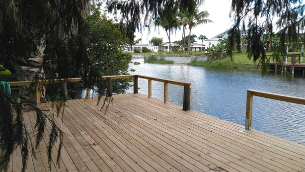 Une terrasse en bois, dotée d’une balustrade, surplombe une étendue d’eau calme, et est entourée de verdure et d’un bâtiment situé au loin.