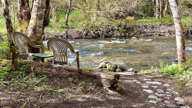Un entorno tranquilo al aire libre con un arroyo cristalino, dos sillas de plástico y un sendero de piedra.