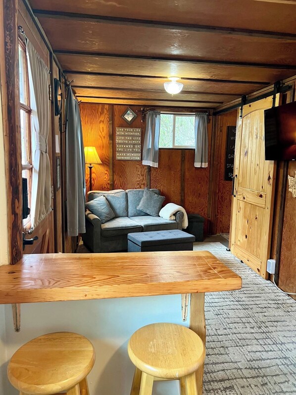Acogedor interior de cabaña con techo de madera, un sofá, una mesa y dos taburetes.