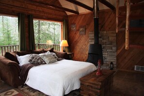 A cozy cabin bedroom with a bed, pillows, a lamp, a fireplace, and a view of the forest.