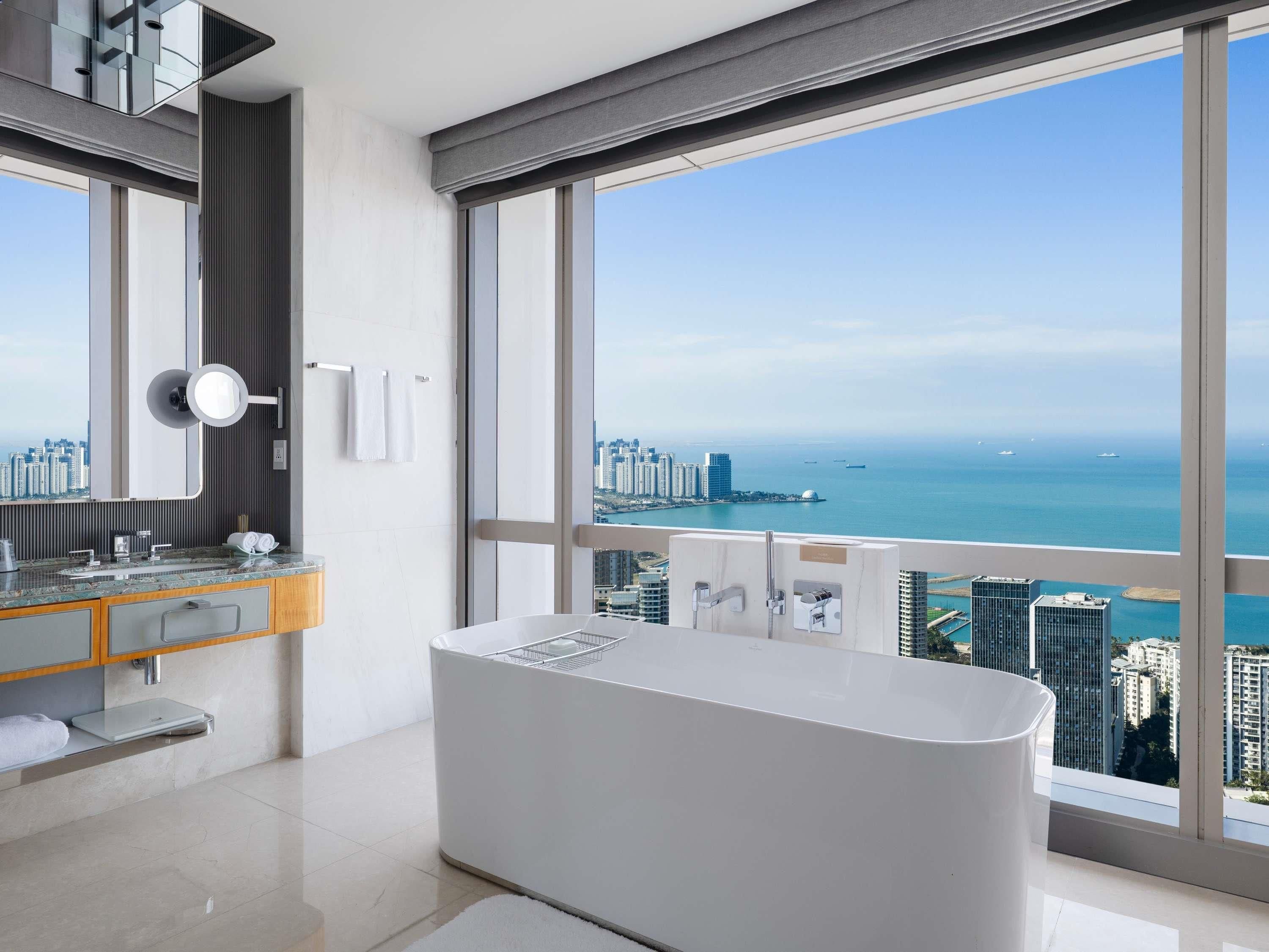 A modern bathroom with a large bathtub, a vanity with a mirror, and a view of the city and sea.