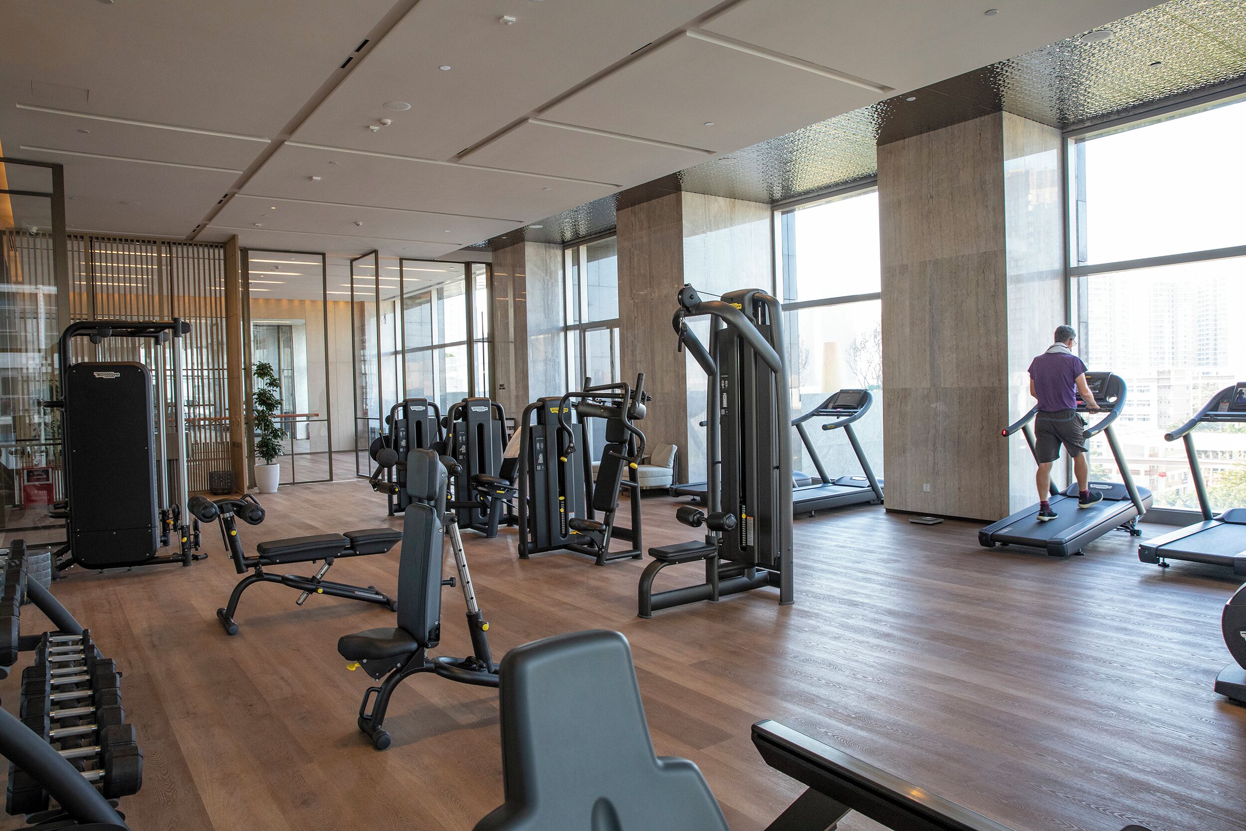 Un gimnasio con diversos equipos de ejercicio, ventanas amplias y piso de madera.