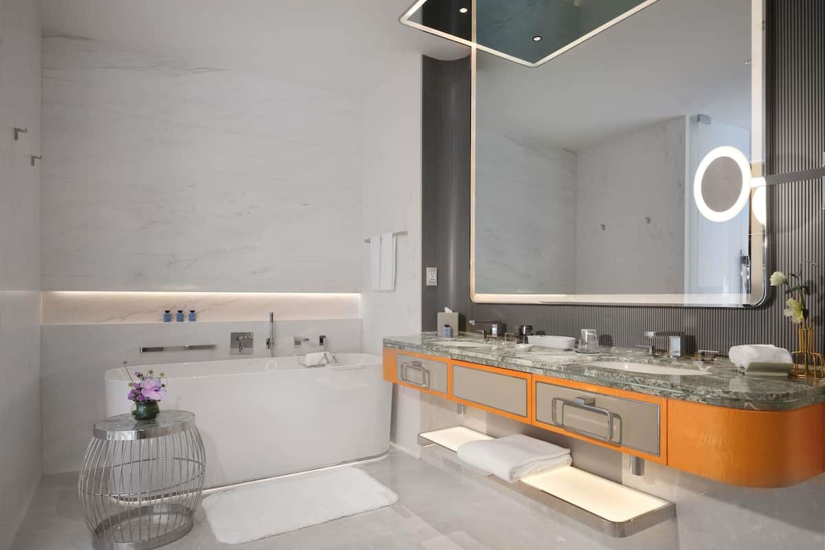 A modern bathroom with a large mirror, a bathtub, a vanity with a sink, and a small round table with a vase.