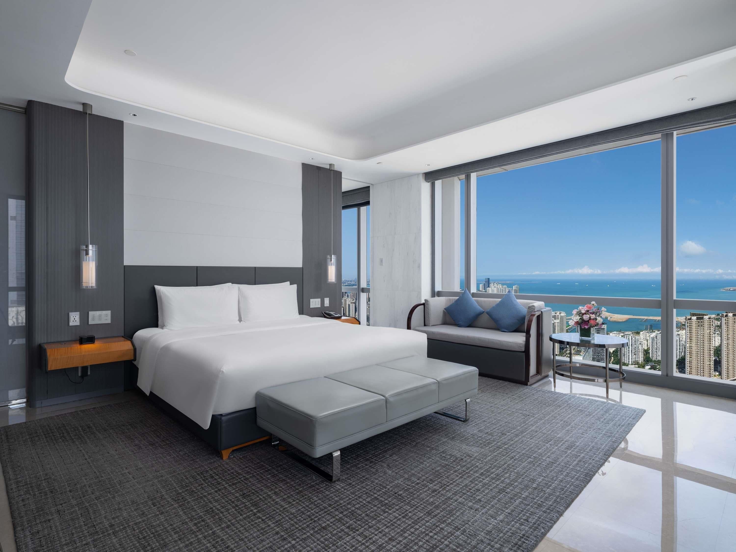 A modern hotel room with a large bed, a sofa, a small table, and a view of the city and ocean.