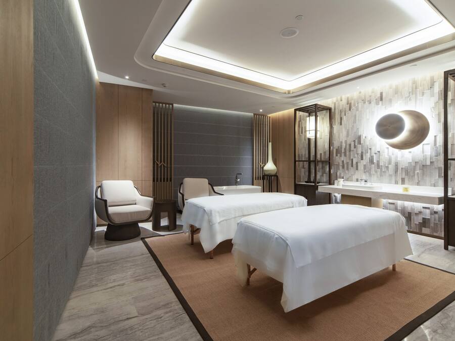 A modern hotel room with two beds, a sitting area, a large mirror, and a bathroom with a bathtub and sink.