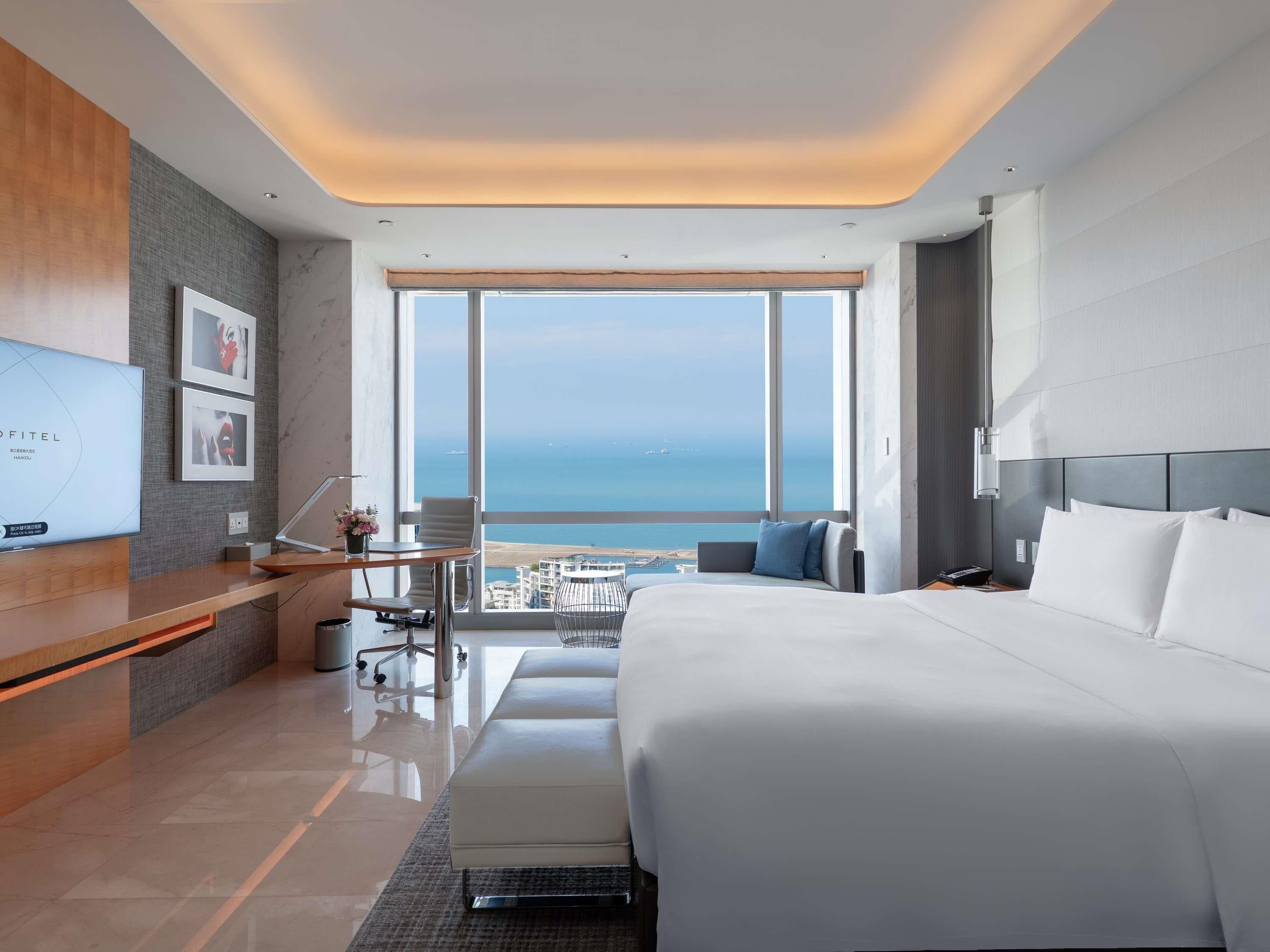 Quarto de hotel moderno com uma cama grande, uma mesa com computador, uma cadeira e vista para o mar.