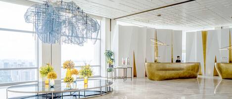 A modern hotel lobby with a glass reception desk, a large abstract light fixture, and a view of the city.