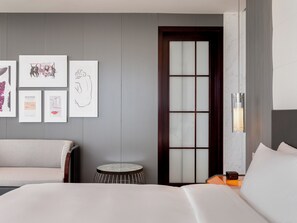 A modern hotel room with a large bed, a round side table, and a sofa. There are framed artworks on the wall and a large window with a wooden door.