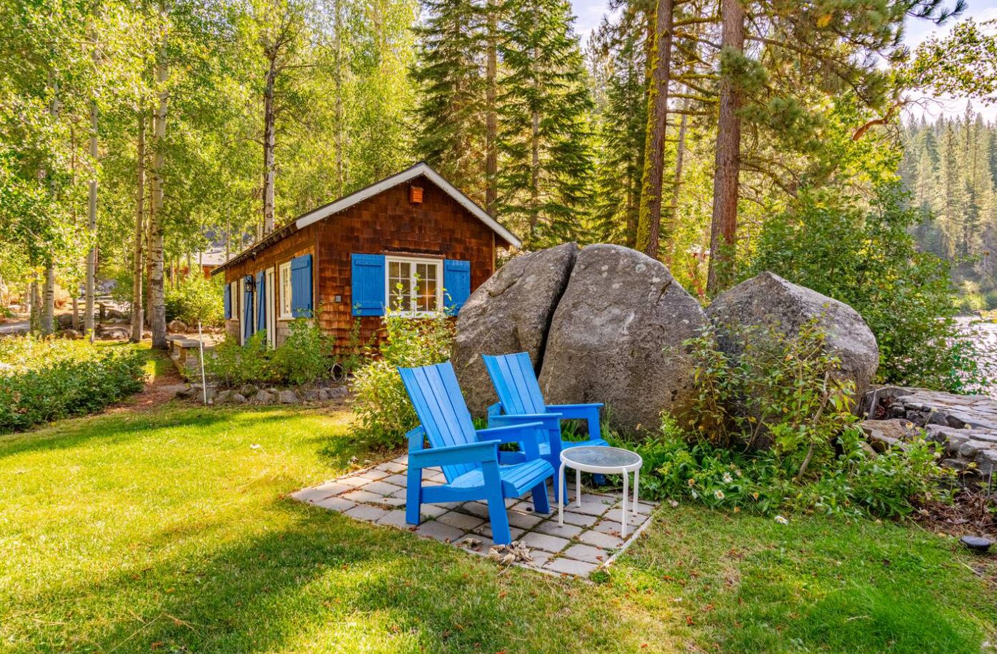 Una cabaña con base de piedra, contraventanas azules y un pequeño porche con dos sillas Adirondack azules y una mesa redonda.