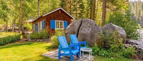 Una cabaña con base de piedra, contraventanas azules y un pequeño porche con dos sillas Adirondack azules y una mesa redonda.