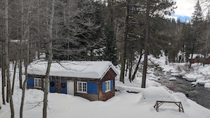 A cabin with a snow-covered roof, surrounded by trees and a snowbank.