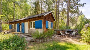 A rustic cabin with a stone foundation, wooden siding, and blue shutters, surrounded by a forested area with a patio and outdoor seating.
