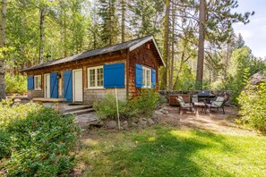 Une cabane rustique avec des fondations en pierre, un revêtement en bois et des volets bleus, entourée d’une zone boisée et dotée d’une terrasse et de meubles d’extérieur.