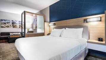 A modern hotel room with a large bed, a wooden headboard, a nightstand, a mirror, and a wall-mounted light.