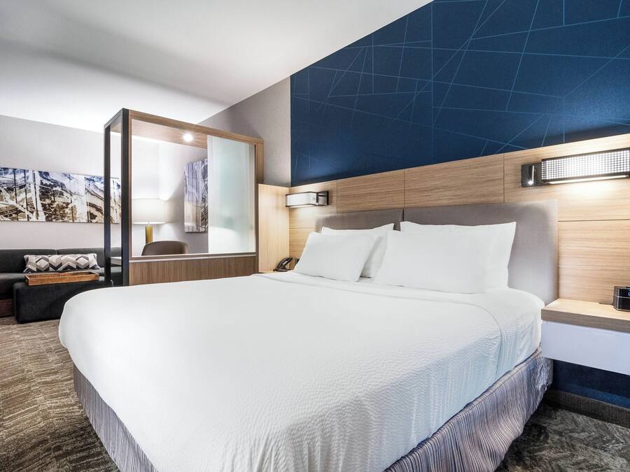 A modern hotel room with a large bed, a wooden headboard, a nightstand, a mirror, and a wall-mounted light.
