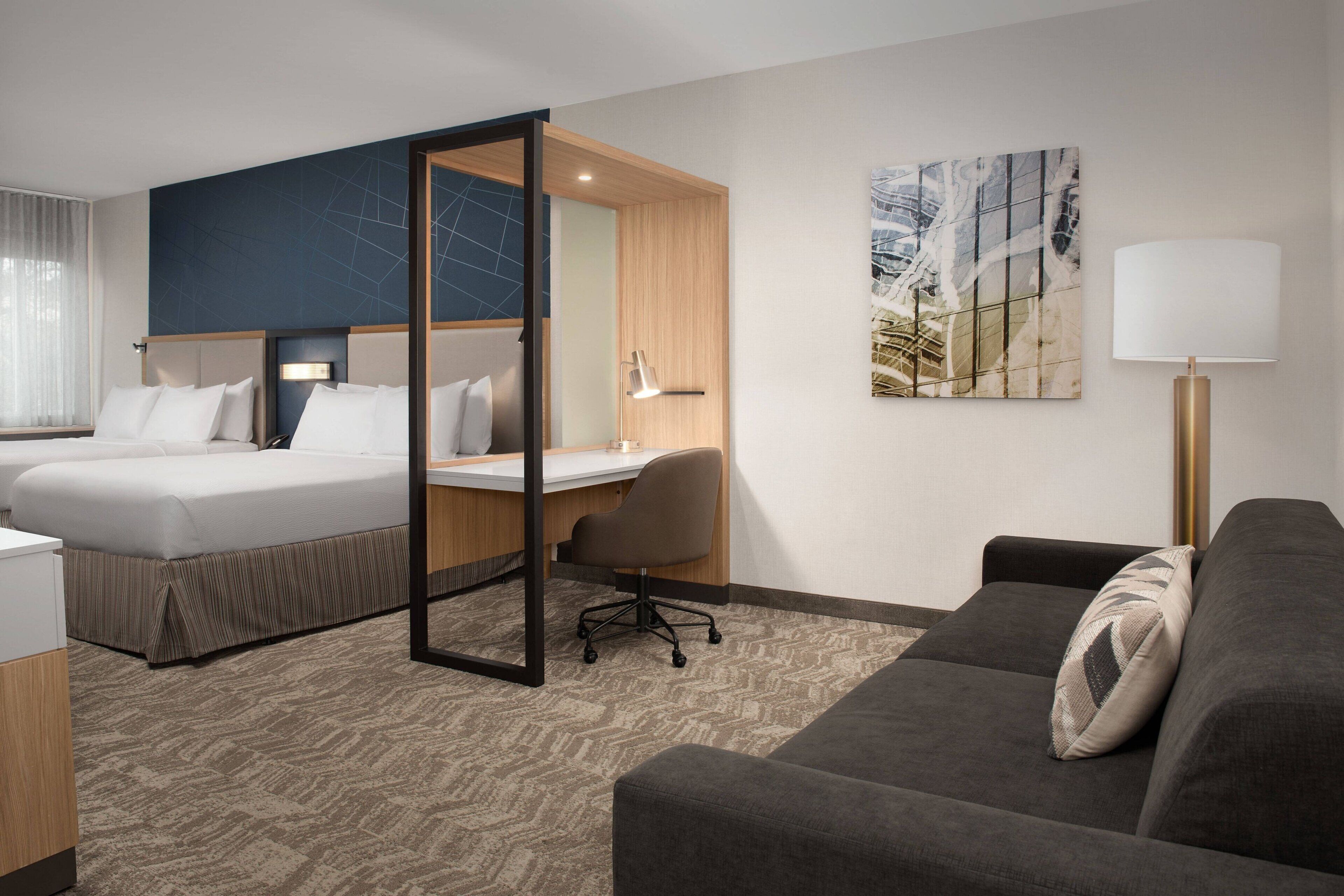A hotel room with two beds, a desk, a chair, a sofa, and a lamp.