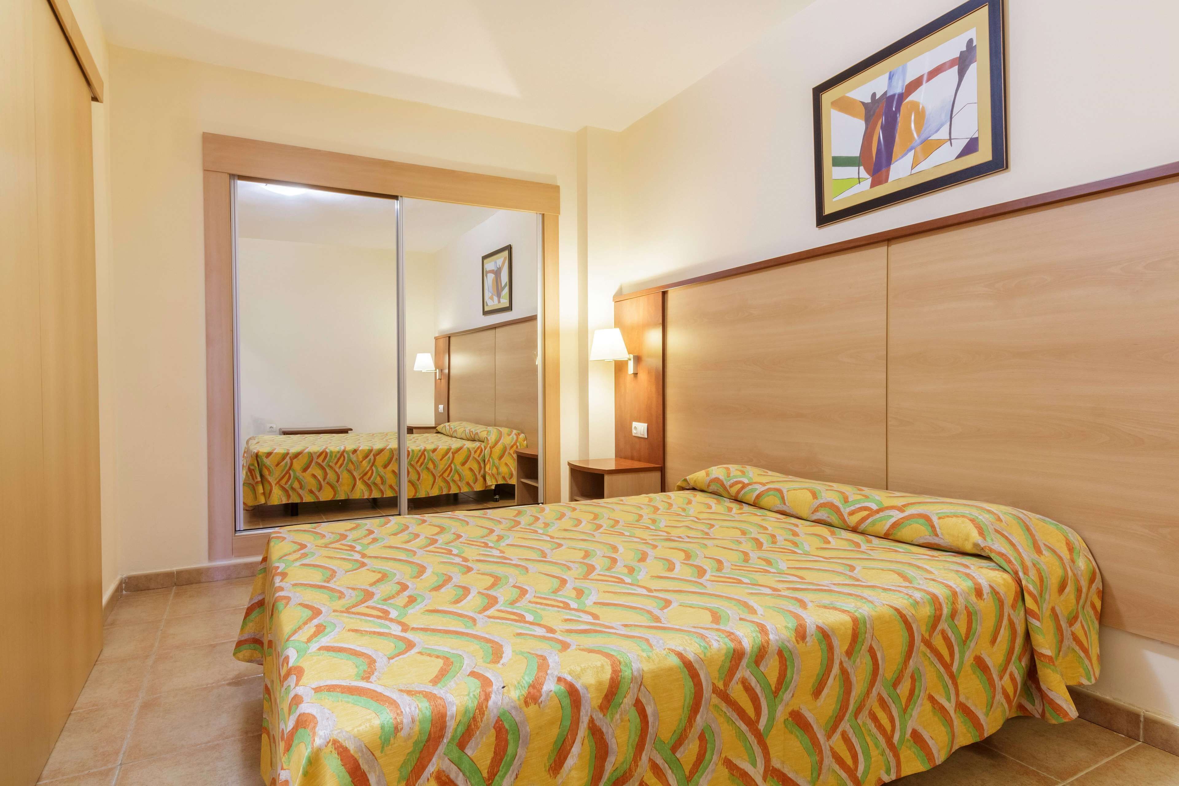 Habitación de hotel con una cama, un espejo, una mesita de noche y una lámpara de pared.