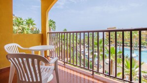 Un balcón con dos sillas de plástico blanco y una mesa redonda pequeña, con vistas a una zona de piscina con palmeras y un edificio al fondo.