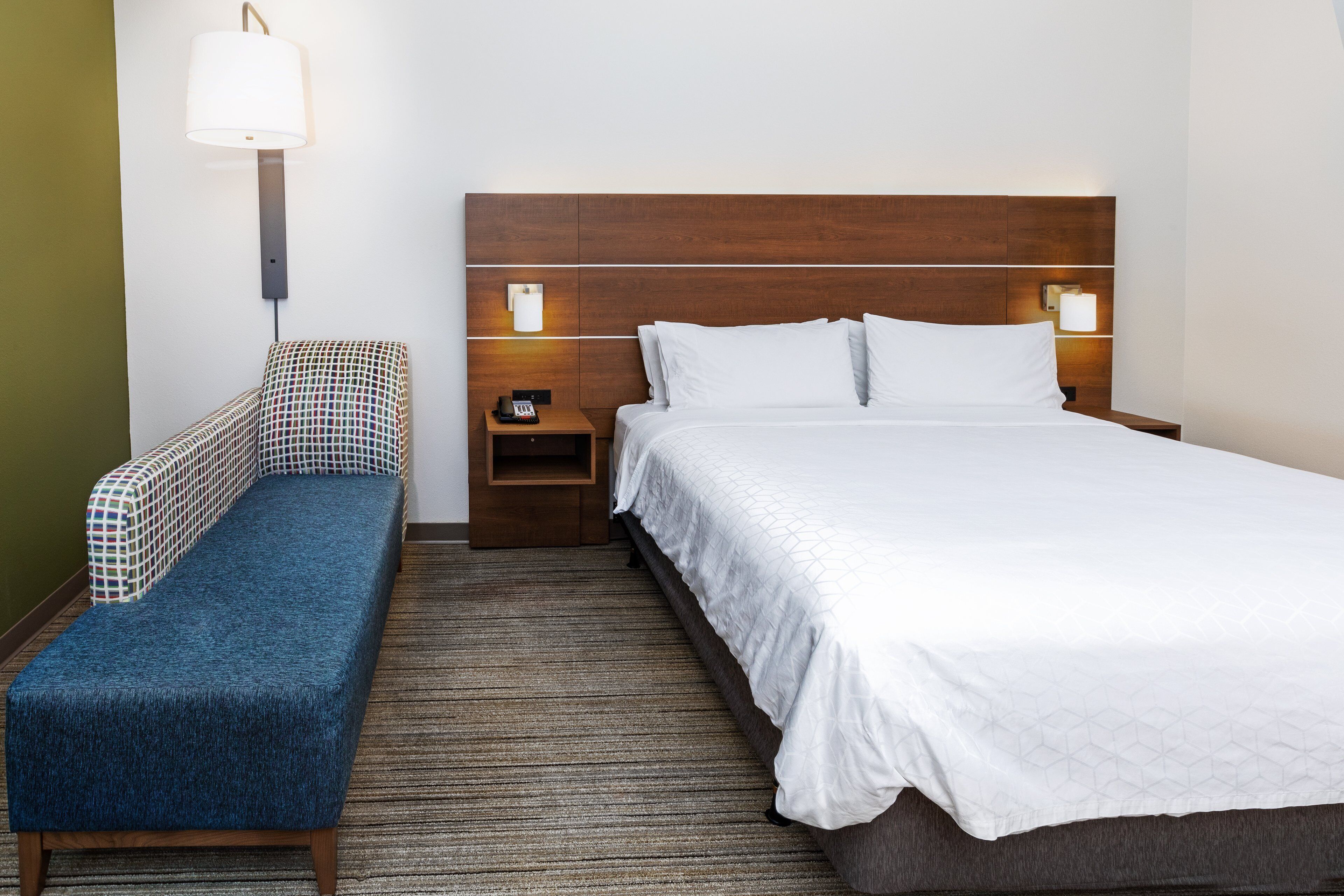A hotel room with a bed, a bench, a nightstand, and a wall-mounted lamp.