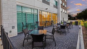 An outdoor seating area with tables and chairs, a brick floor, and a building with large windows and a glass door.