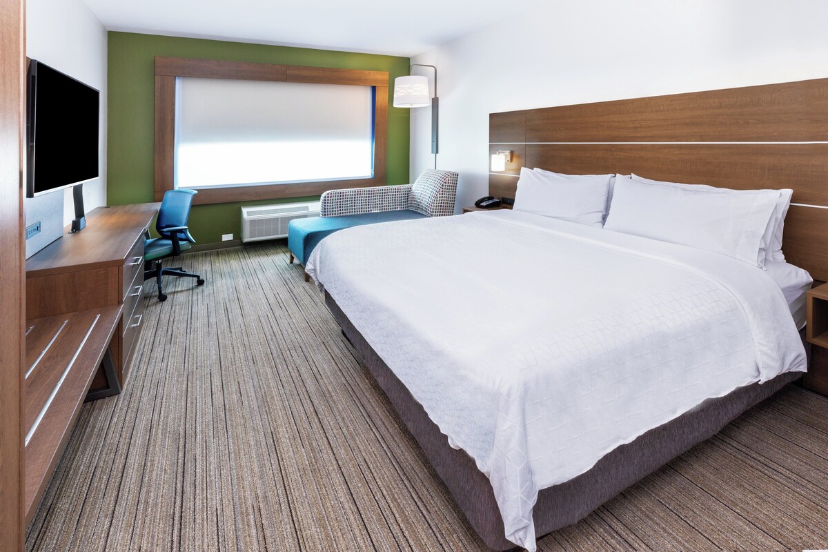 Quarto de hotel com uma cama grande, uma escrivaninha com televisão de tela plana, uma cadeira e uma janela com persianas.