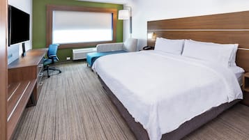 Ein Hotelzimmer mit einem großen Bett, einem Schreibtisch mit Computer, einem Sessel, einer Couch und einem Fenster mit Jalousien.
