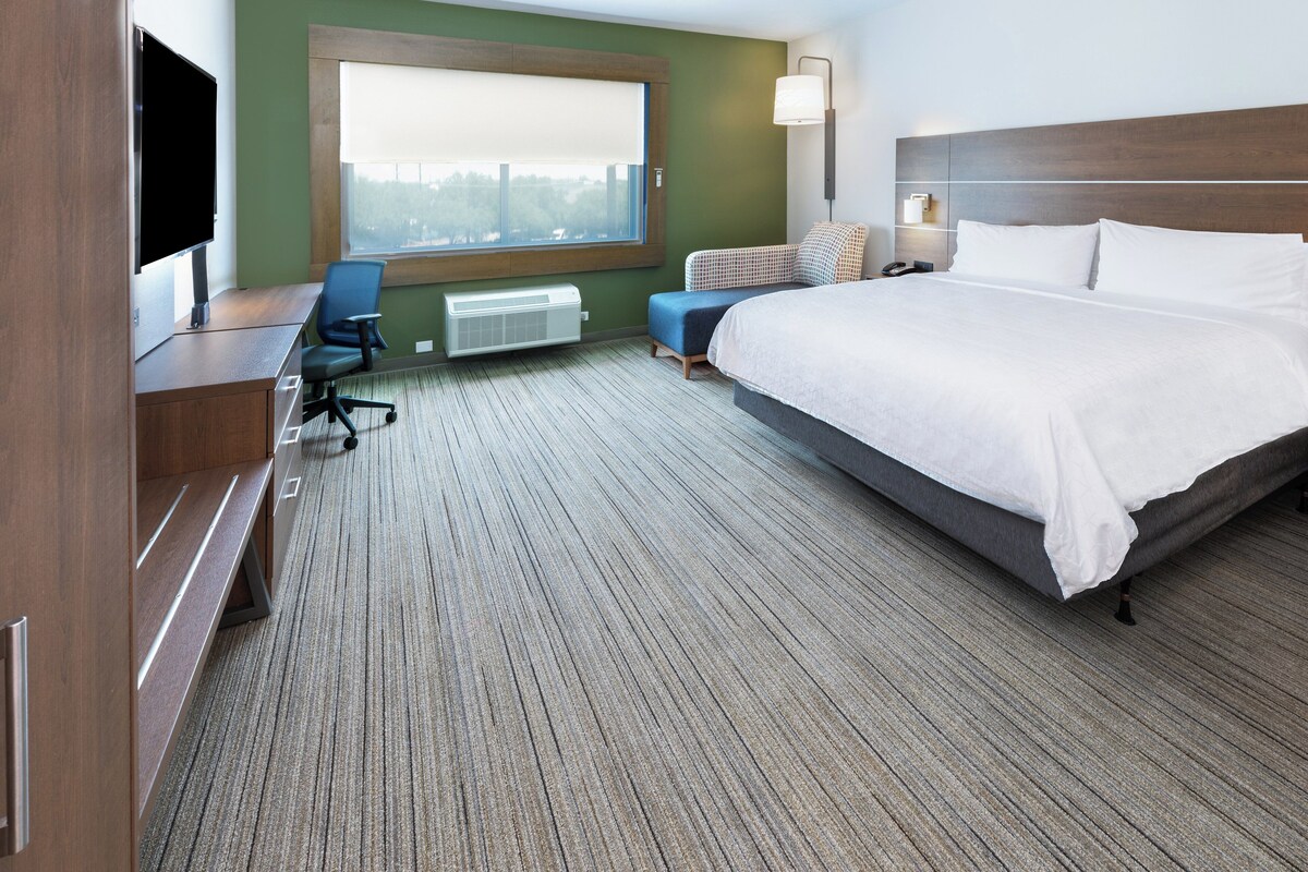 Quarto de hotel com uma cama grande, uma escrivaninha com cadeira, uma televisão, uma janela com persianas e um abajur.