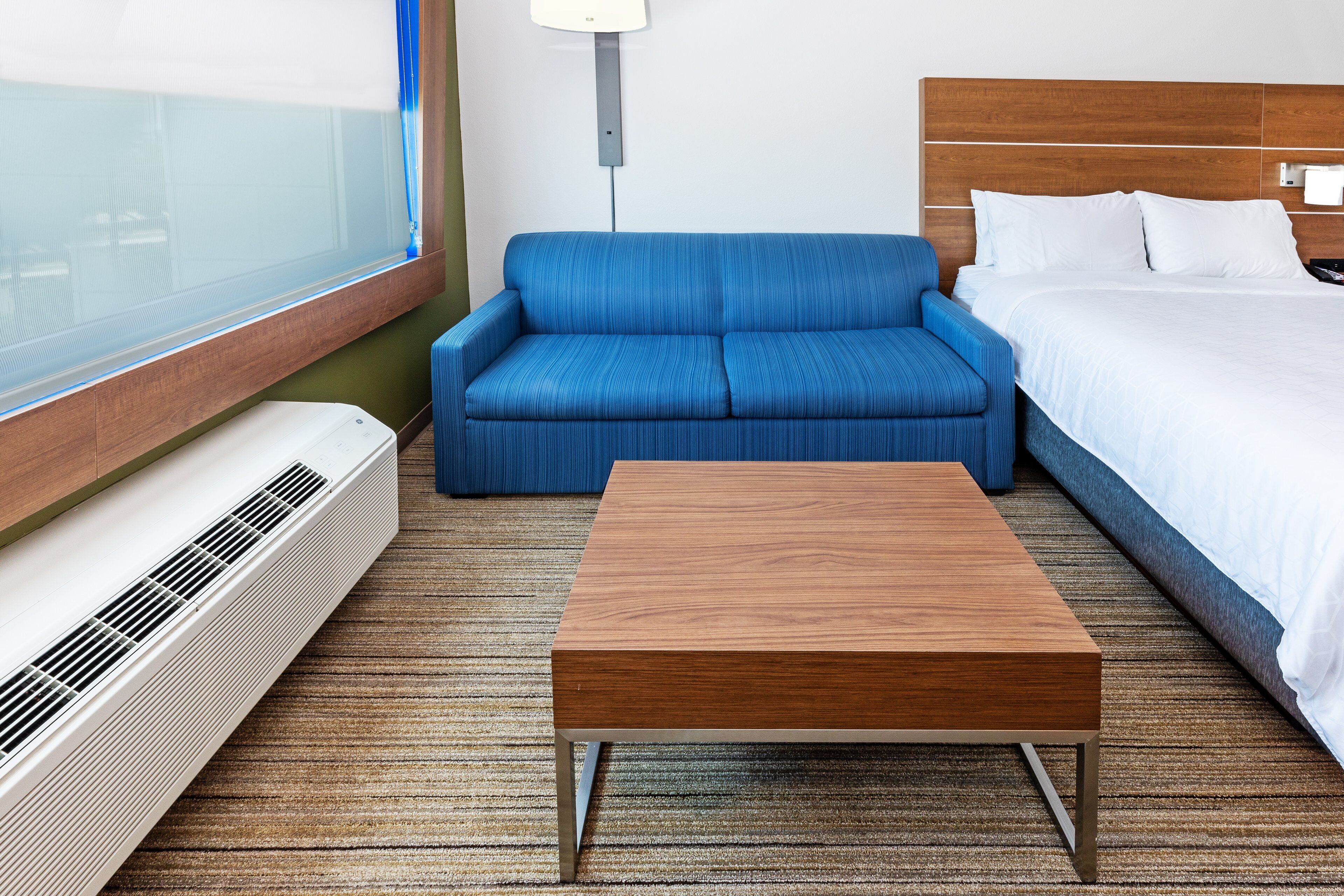 A hotel room with a blue sofa, a wooden coffee table, a bed with white linens, and a wall-mounted heater.