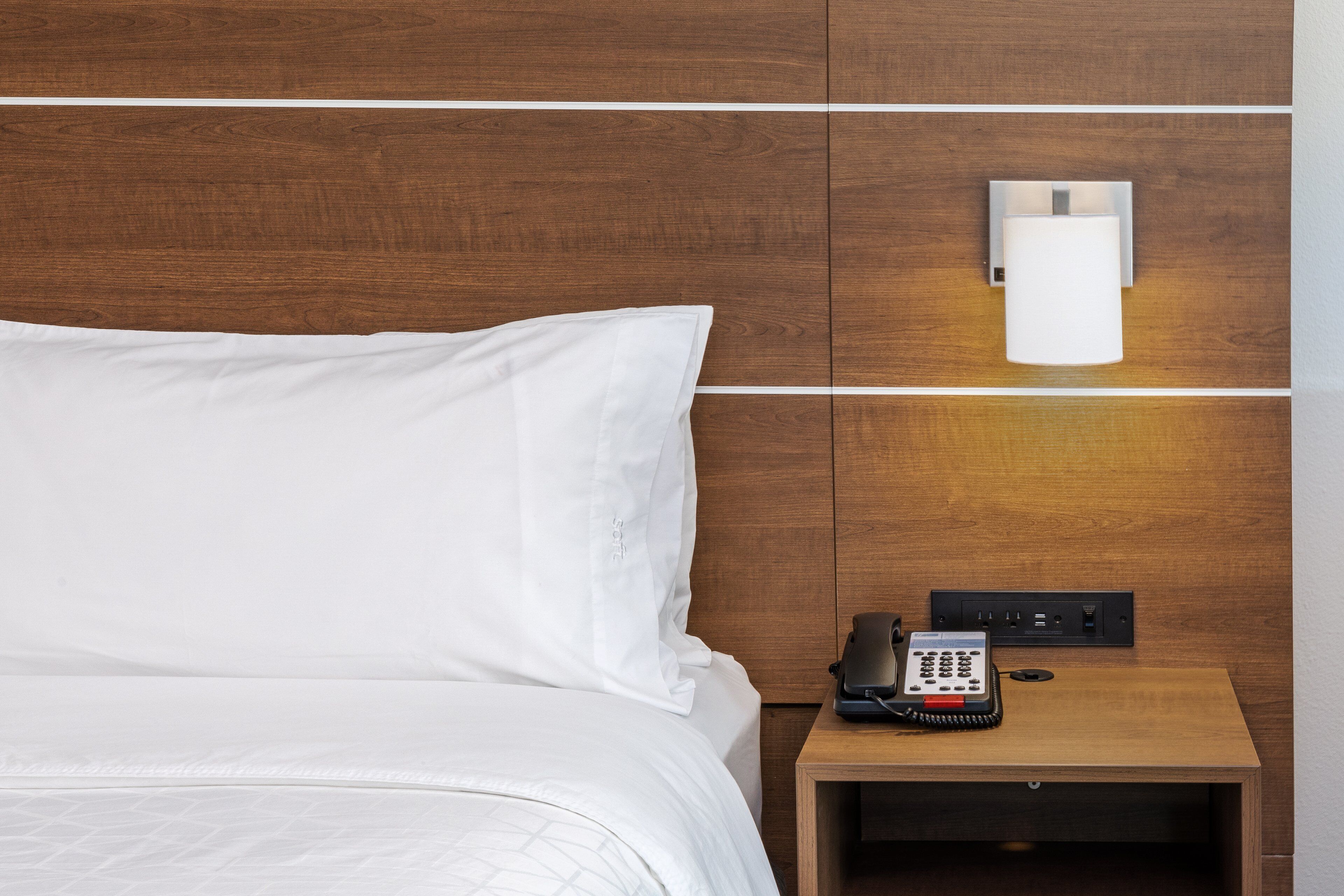 Een keurig opgemaakt bed met witte lakens, een nachtkastje met telefoon en stekkerdoos, en een wandlamp.