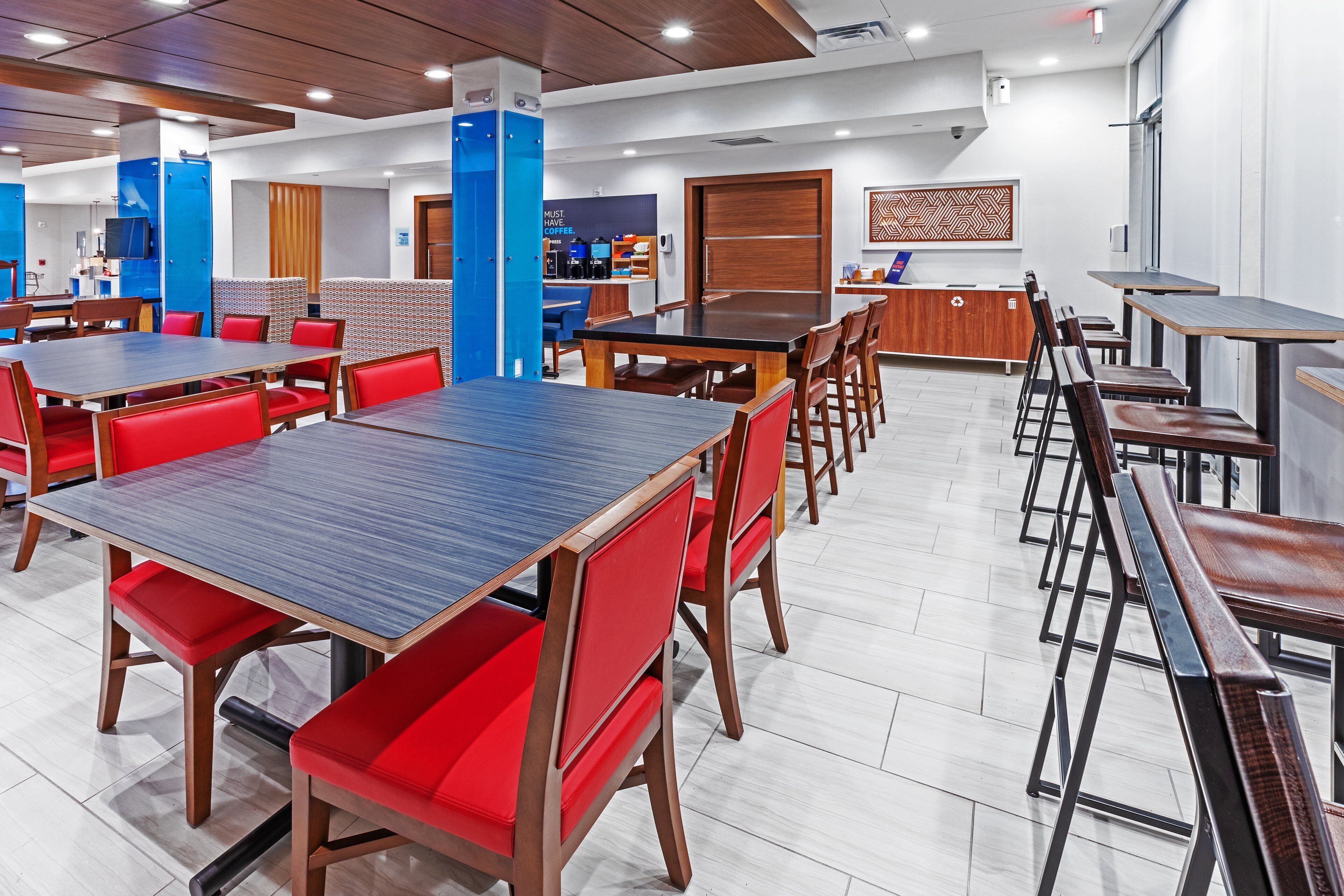 A modern dining area with multiple tables and red chairs, a coffee station, and a counter with a menu display.