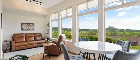 Un vaste espace de vie doté d’une grande fenêtre offrant une vue panoramique, d’un canapé en cuir marron, d’une table de salle à manger ronde avec des chaises et d’un ventilateur de plafond.