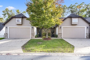 A two-story house with a central tree, two garage doors, and a well-maintained lawn.