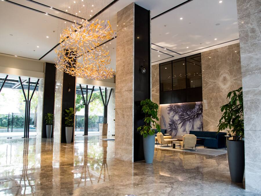 A modern lobby with a marble floor, large potted plants, a unique ceiling light fixture, and a seating area with a blue sofa and chairs.