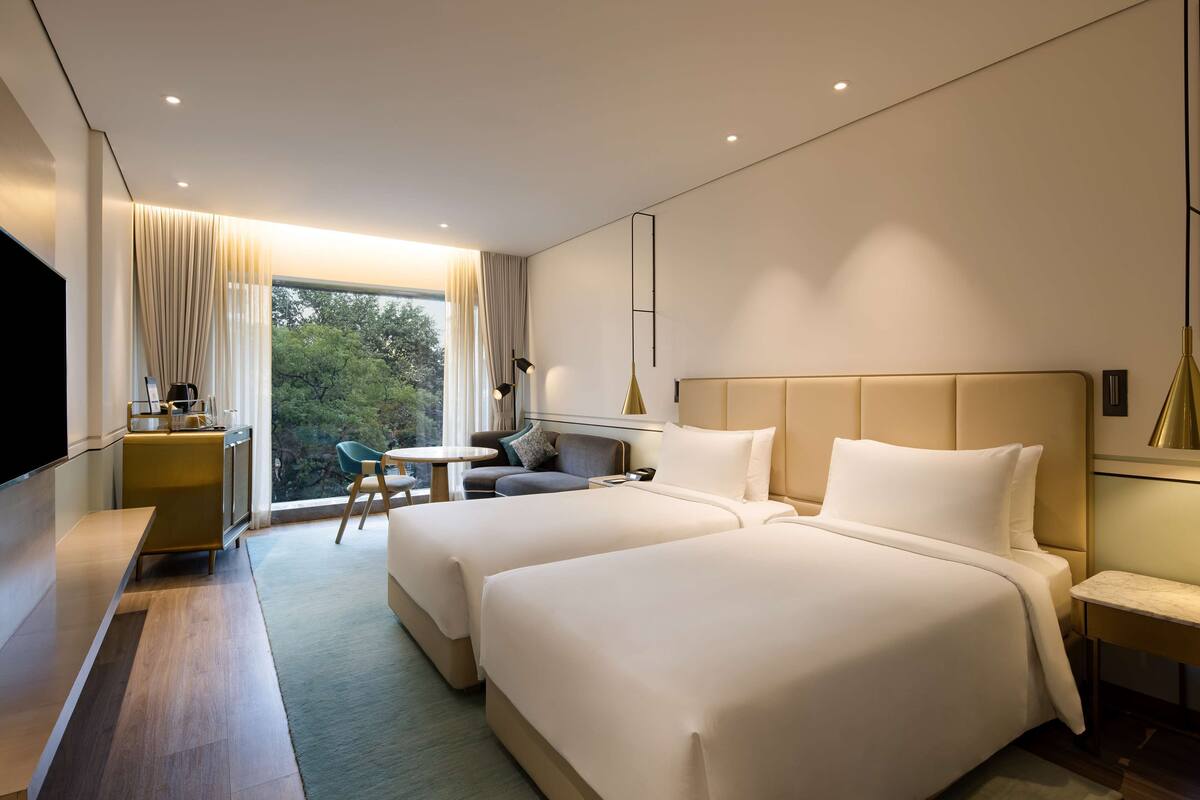 A modern hotel room with two beds, a desk, a chair, and a large window offering a view of trees.