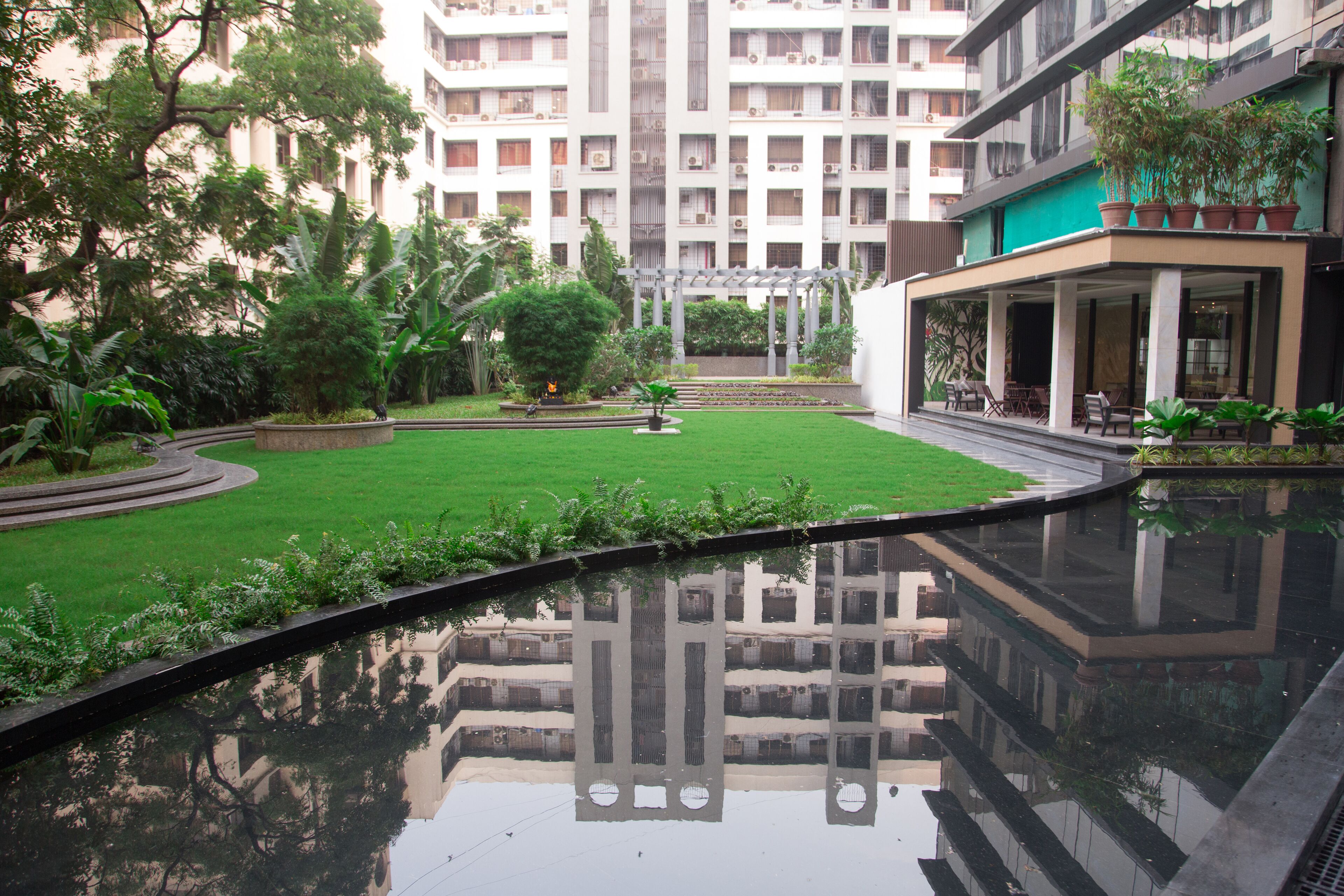 A courtyard with a reflective pool, lush greenery, and a multi-story building in the background.