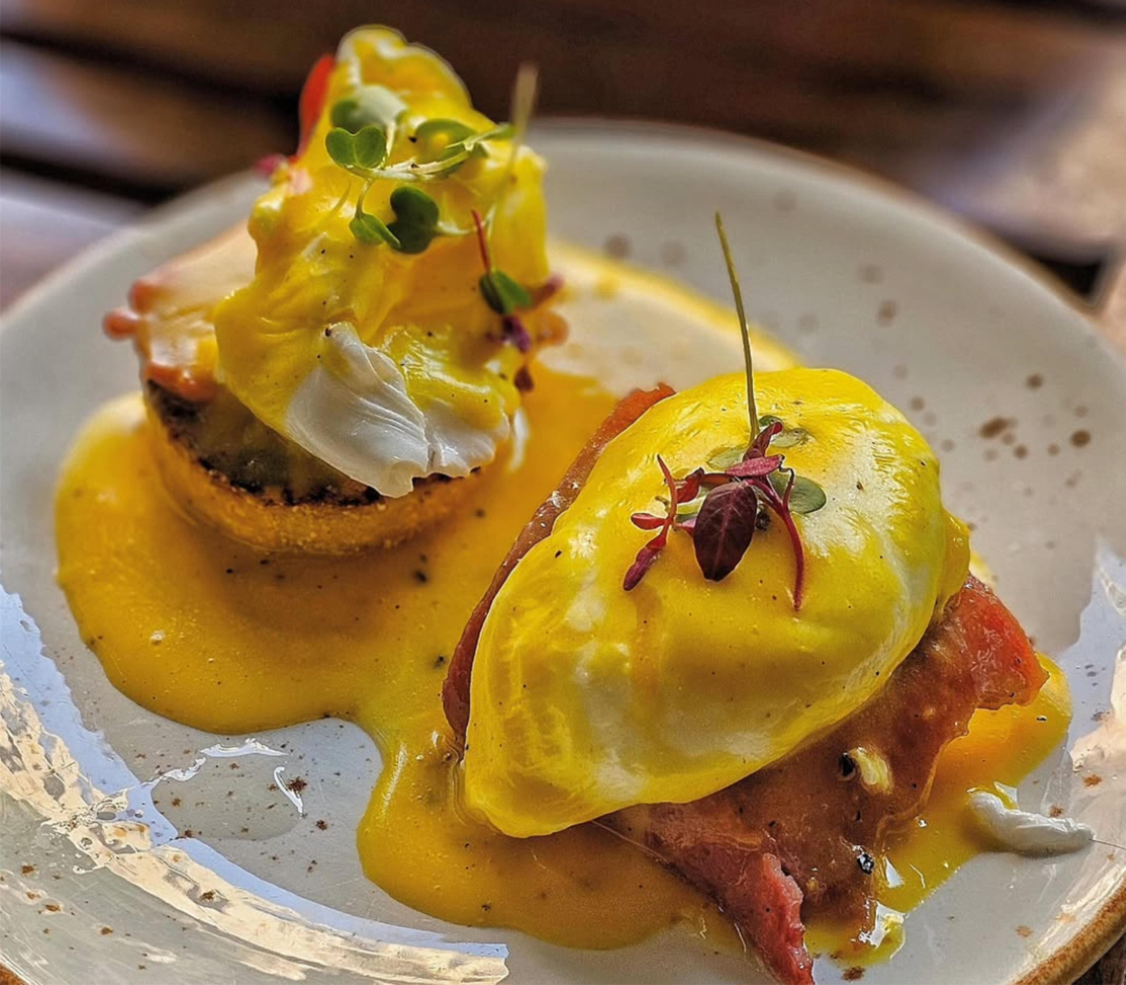 Een bord met twee Eggs Benedict gerechten, elk overgoten met hollandaisesaus en versierd met microgroenten.