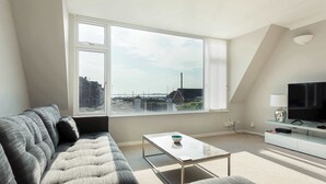A modern living room with a large window, a sofa, a coffee table, and a TV stand. - Reel Time - A Stone's Throw from Poole Quay (Poole)