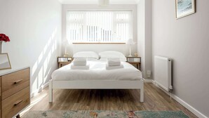 A bedroom with a white bed, wooden floor, a radiator, a nightstand with a lamp, and a framed picture on the wall. - Reel Time - A Stone's Throw from Poole Quay (Poole)