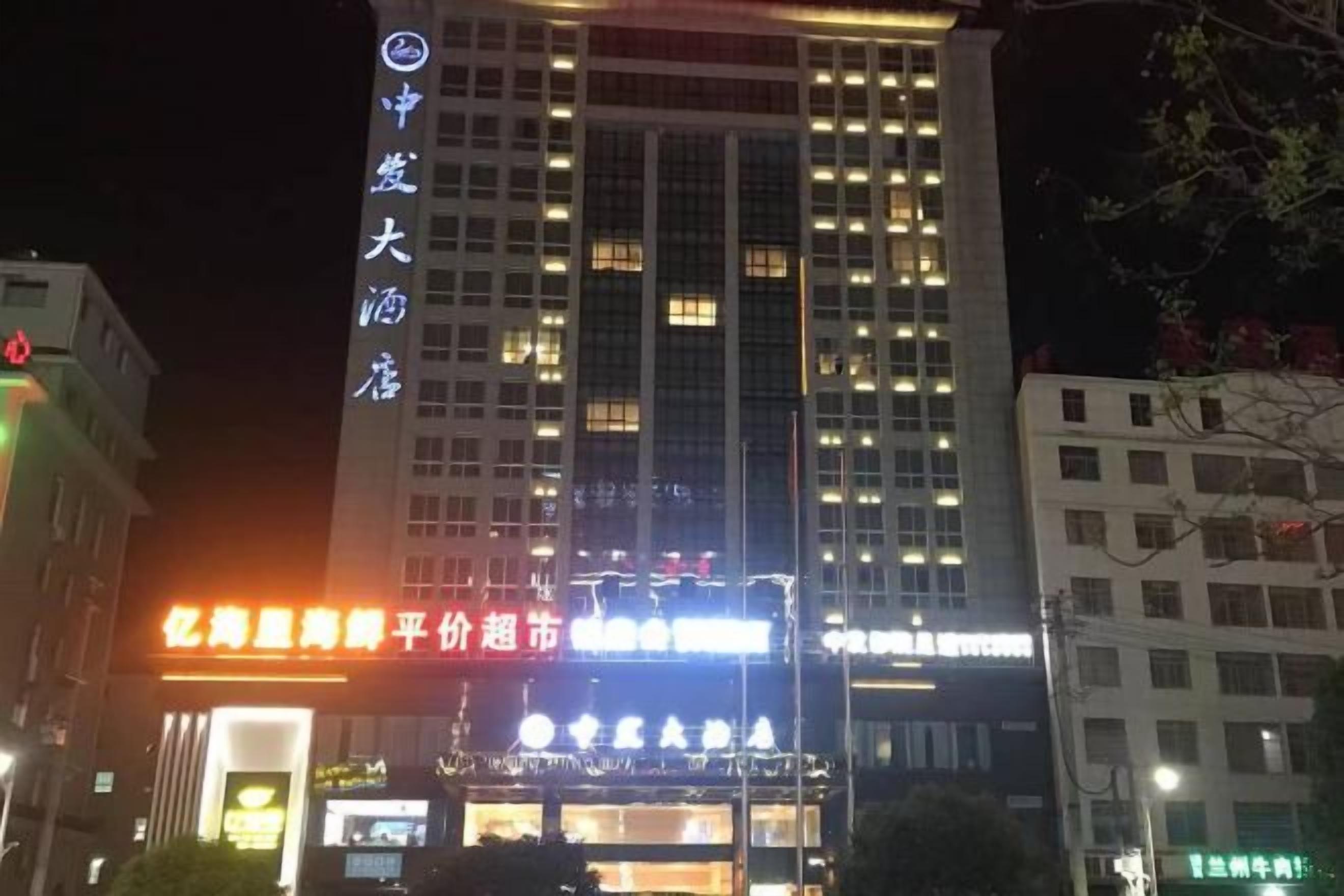 Un hotel de varios pisos con señalización iluminada, un cartel vertical llamativo a la izquierda y una entrada muy iluminada.